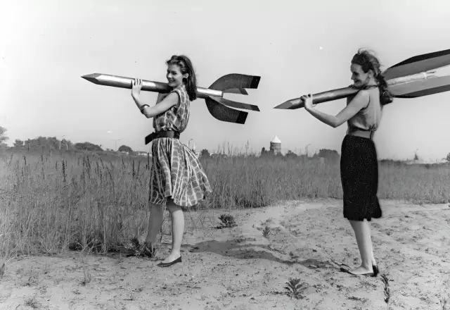 Zwei junge Frauen - Raketenmädchen - tragen Holzraketen auf ihren Schultern. Die schwarz-weiße Aufnahme zeigt sie in einer statischen Pose.