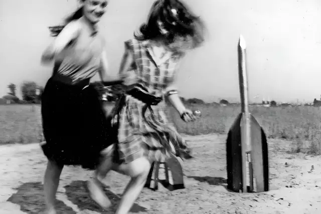 Zwei junge Frauen - Raketenmädchen - vor einer Holzrakete. Die schwarz-weiße Aufnahme zeigt sie in einer dynamischen, laufenden Pose.