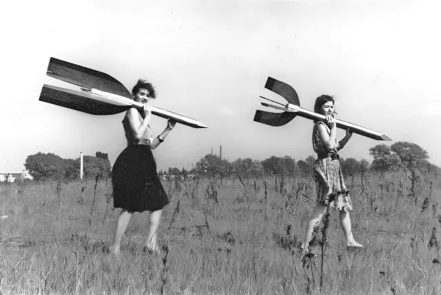 Zwei junge Frauen - Raketenmädchen - tragen Holzraketen auf ihren Schultern. Sie tragen Röcke, im Hintergrund eine karge Landschaft.