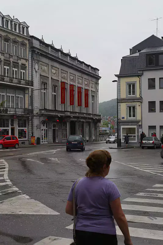 Frau in Rückenasicht an einem trüben Tag in Spa, Belgien. Die Frau trägt einen lila Shirt und überquert eine Kreuzung mit Zebrastreifen